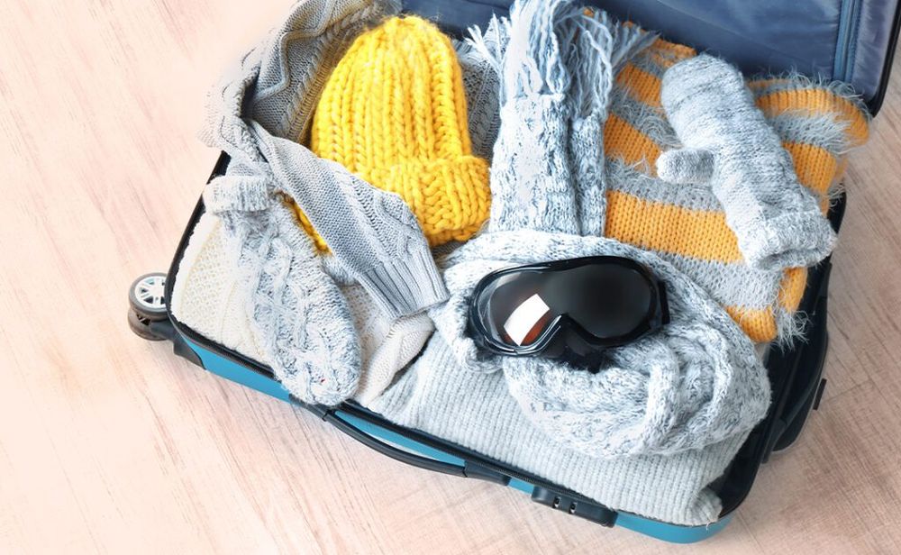 Open suitcase with warm clothes and ski goggles on wooden floor.